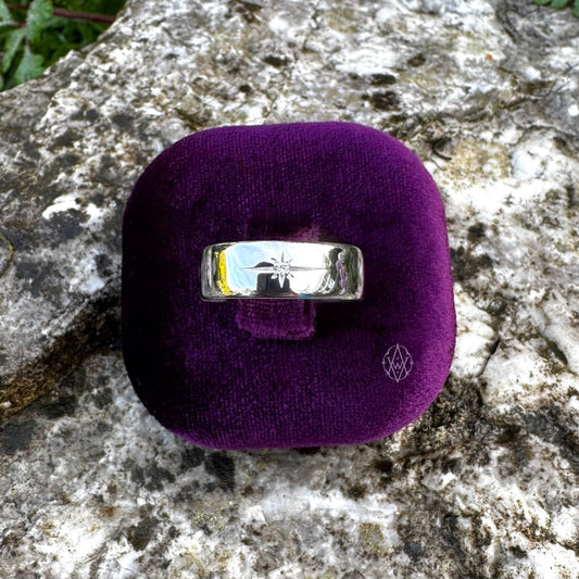A polished silver band ring with a starburst engraving, displayed on a small purple velvet ring box set against a textured gray stone background.