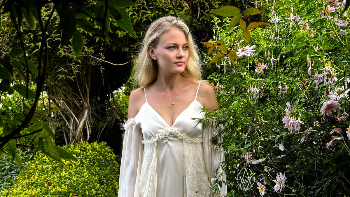 Ashlen, blonde woman in white dress, wearing pendant and earrings, stands in garden with trellis and flowers.