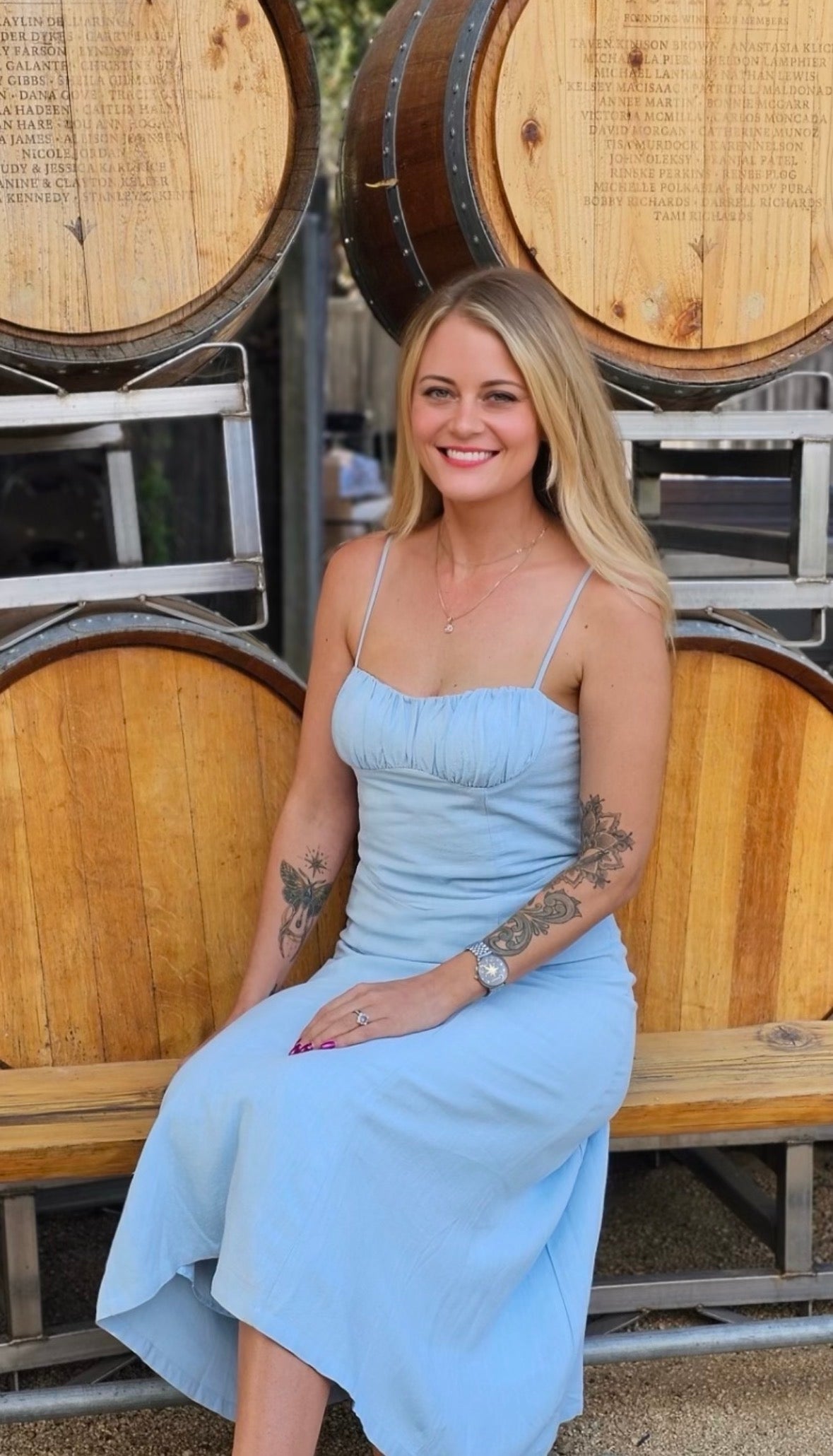 A woman, Ashlen the owner, with blonde hair and tattoos sits on a wooden bench in front of large wooden wine barrels, wearing a light blue sleeveless midi dress. Photo taken in Carmel, CA by Aimee Stevens.