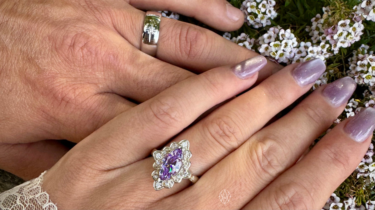 Two hands cross over each other, one on top of the other. The top hand has a purple marquise engagement ring. The bottom hand has a traditional style silver wedding band. There are small white flowers in the background. Photo taken in Roche Harbor by Ashlen VanDeBogart.
