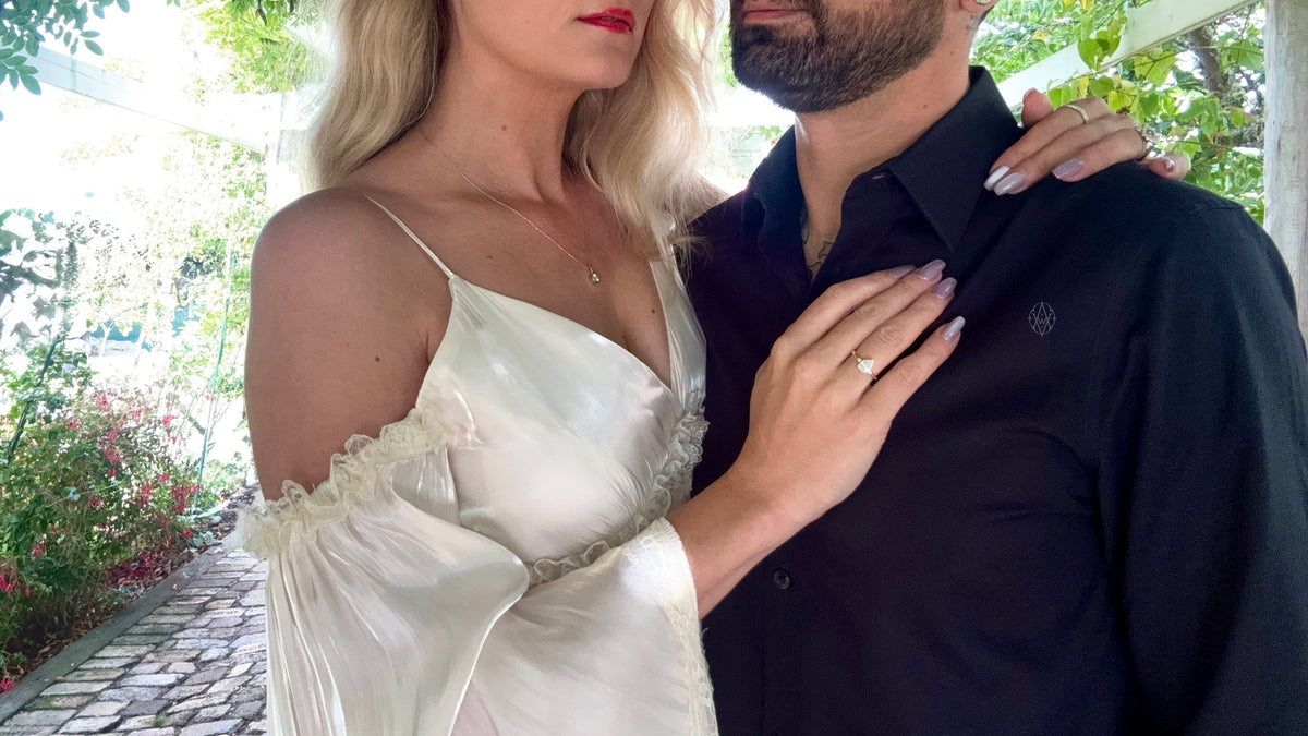 A blonde woman in a white satin slip dress with ruffled trim stands close to a man wearing a black button-up shirt. Her hand rests on his chest, and the Moondrop ring is visible on one finger. She wears a simple necklace.