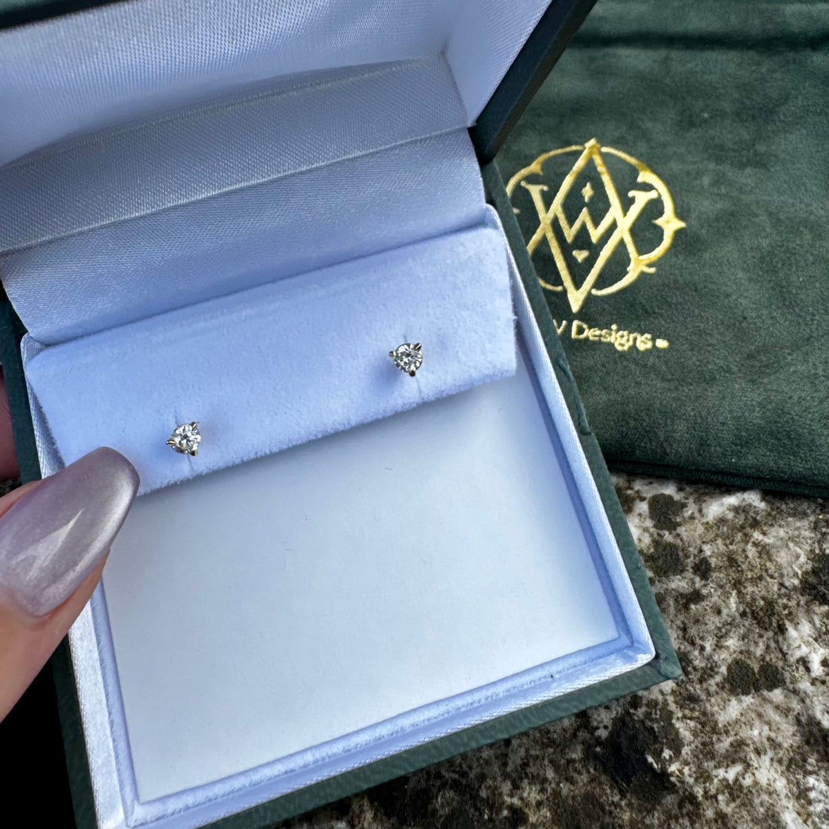 A pair of small round-shaped stud earrings with clear stones in a silver setting, displayed inside an open jewelry box. A hand with metallic lavender nail polish holds the left side of the box. A green logo gift pouch is in the background.