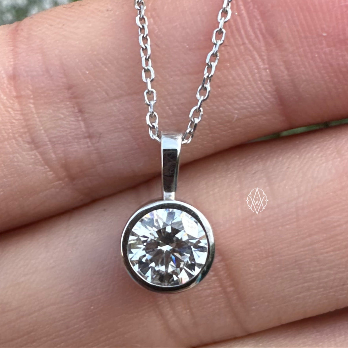 A white gold necklace with a round bezel-set clear diamond pendant, shown resting on a person's hand with blurred flowers in the background.