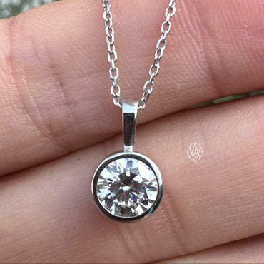 A white gold necklace with a round bezel-set clear diamond pendant, shown resting on a person's hand with blurred flowers in the background.