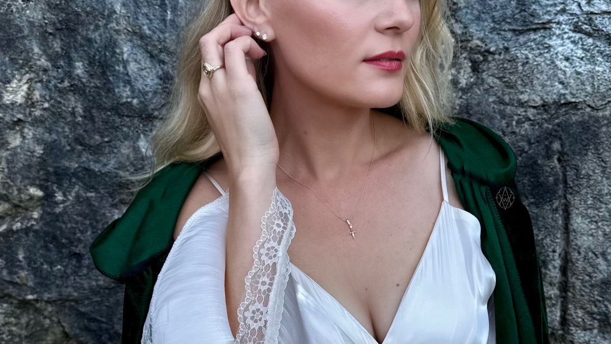 Ashlen, blonde woman in white dress with green cloak, stands in front of stone wall. She wears dagger necklace, stud earrings, and signet ring from the Valor collection. Photo taken in Roche Harbor by Kyle Ketcham.